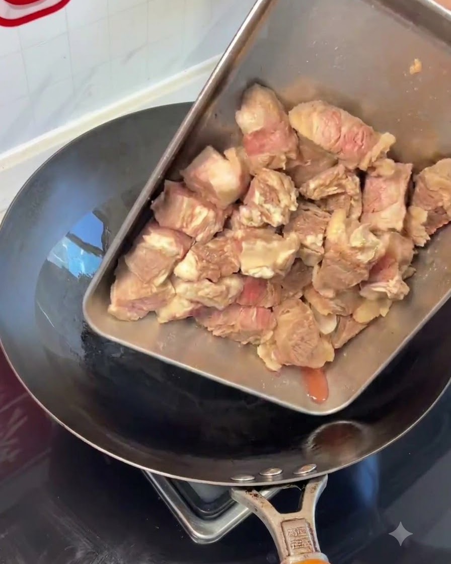 Morceaux de côtes de bœuf blanchies versés depuis un plateau en acier dans un wok chaud.