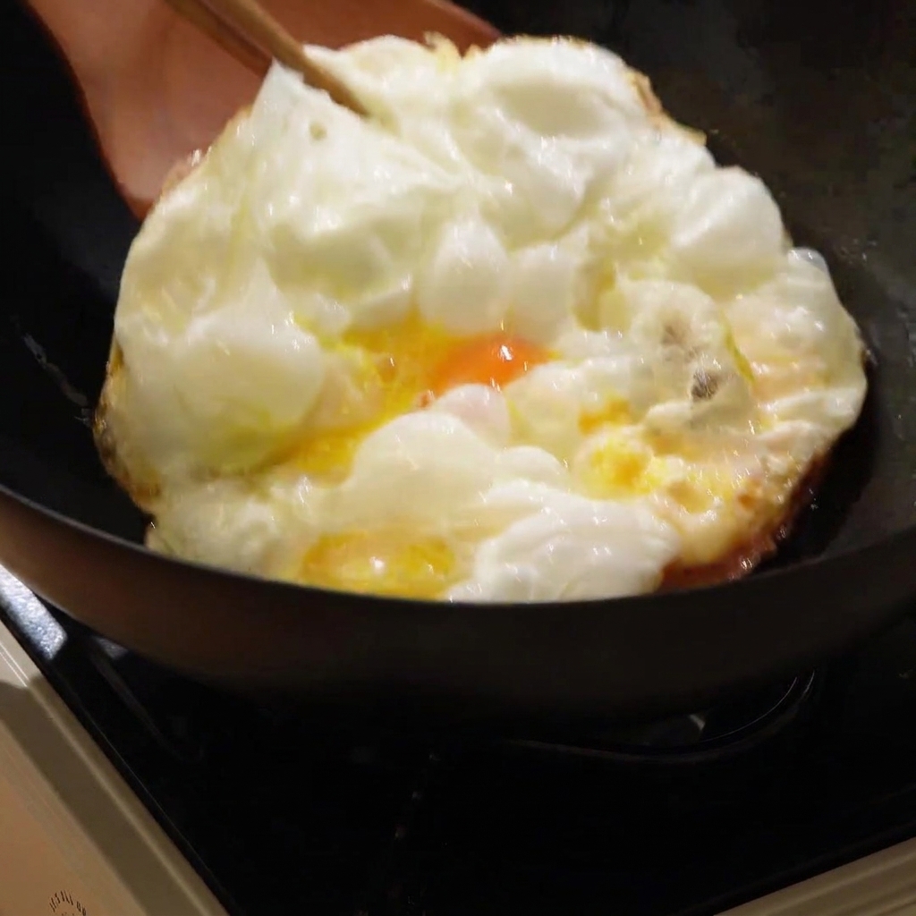 Un grand œuf frit doré en train dêtre retourné dans un wok chaud avec une spatule en bois.