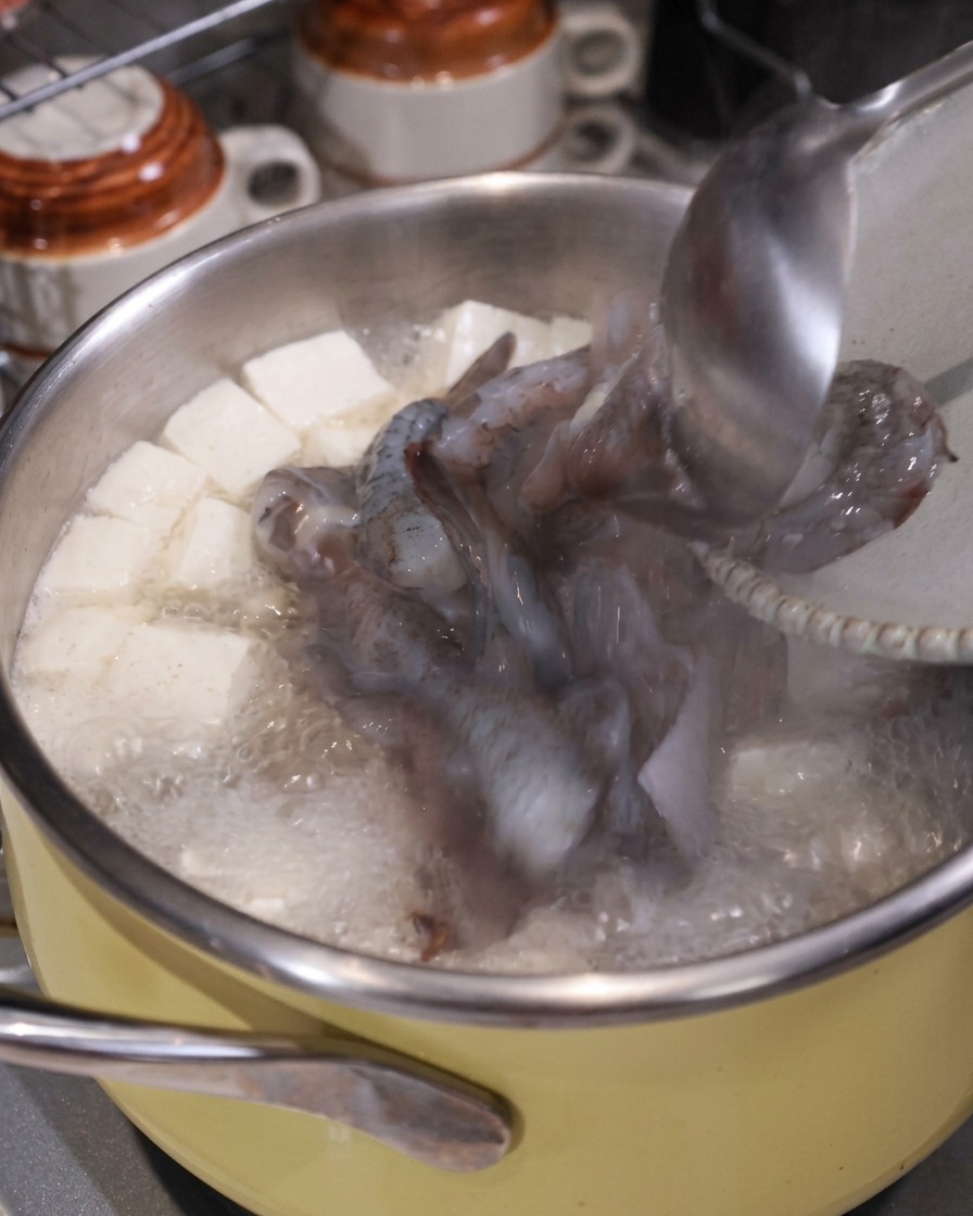 Une louche ajoutant des morceaux de calmar cru et de crevettes dans une casserole de soupe bouillante avec du tofu et du porc.