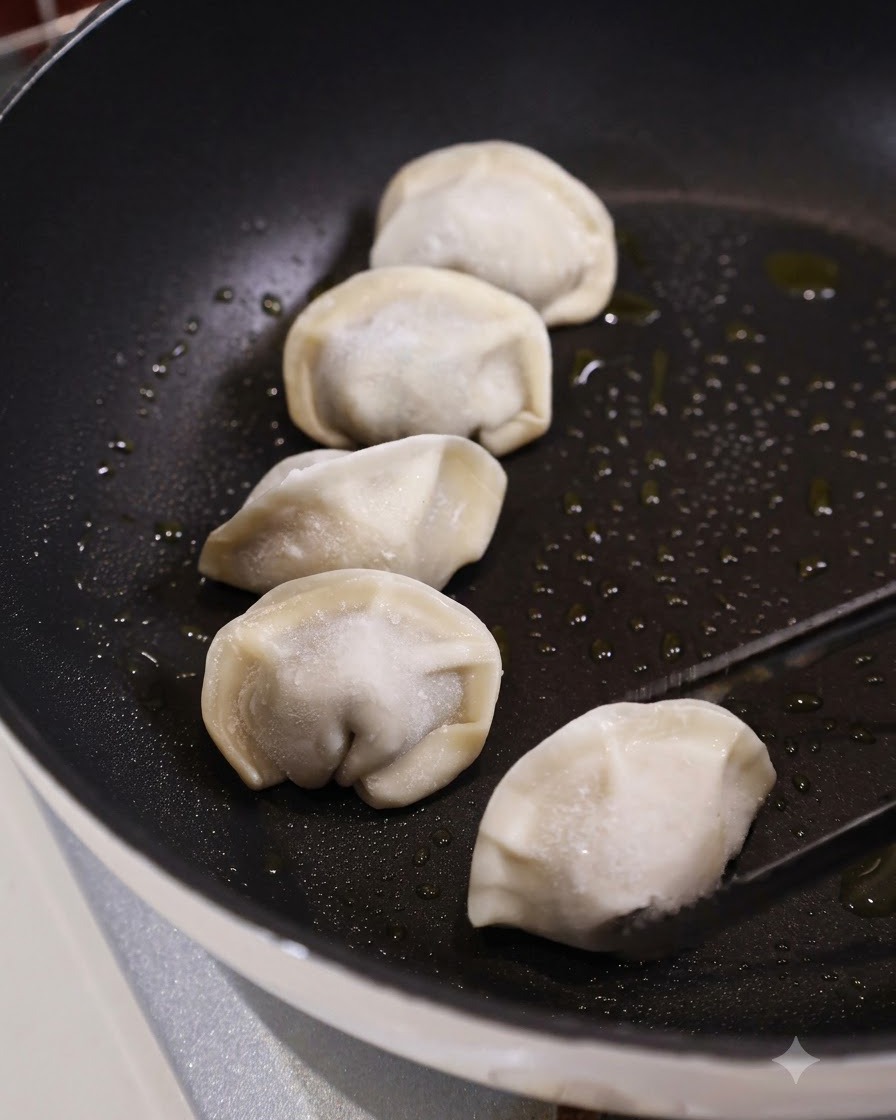 Des pinces de cuisine plaçant un ravioli surgelé cru dans une poêle plate foncée huilée aux côtés de plusieurs autres raviolis.