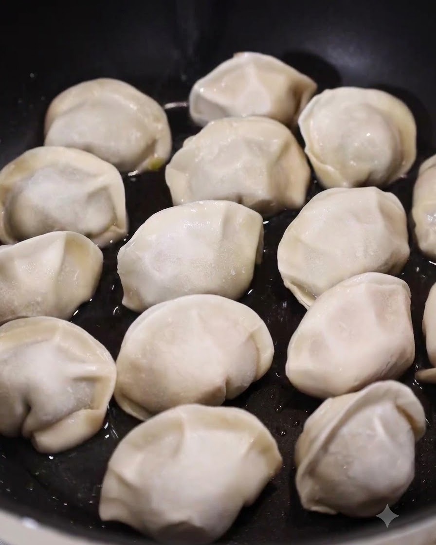 Une poêle plate remplie de raviolis crus reposant dans une fine couche deau et dhuile frémissante.