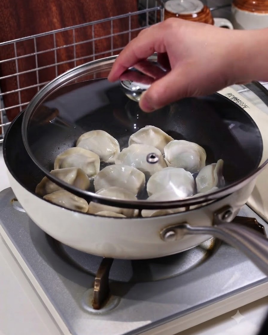 Une main plaçant un couvercle en verre transparent sur une poêle contenant des raviolis fumants sur une cuisinière.