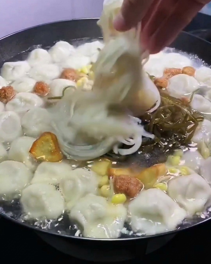 Une main plaçant du varech vert émincé et des nouilles de verre translucides dans une marmite de raviolis et bouillon.