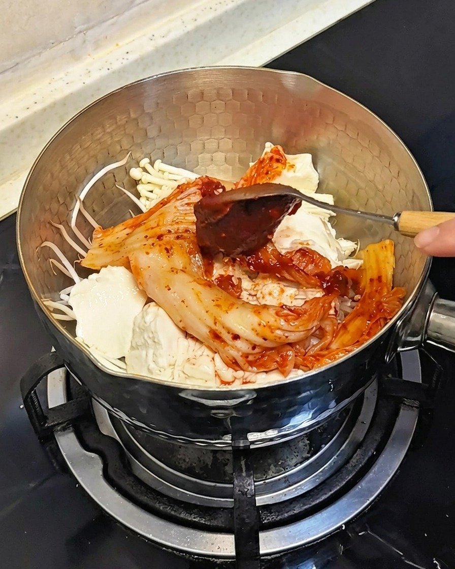 Une cuillère ajoutant une noisette de pâte de piment coréenne rouge et épaisse sur les ingrédients de la casserole.