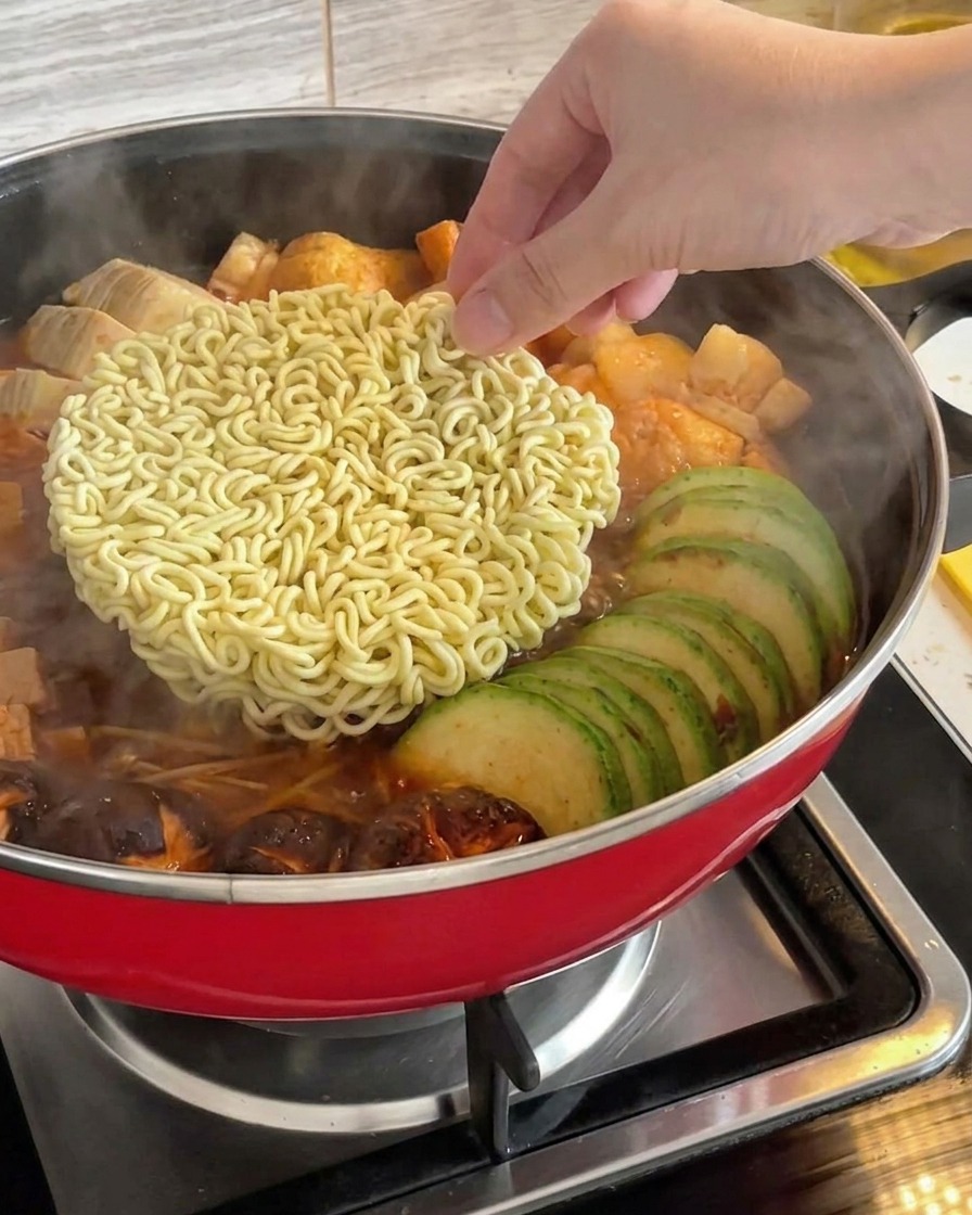 Une main plaçant un bloc de nouilles ramen instantanées sèches au centre dun ragoût de larmée coréenne frémissant.