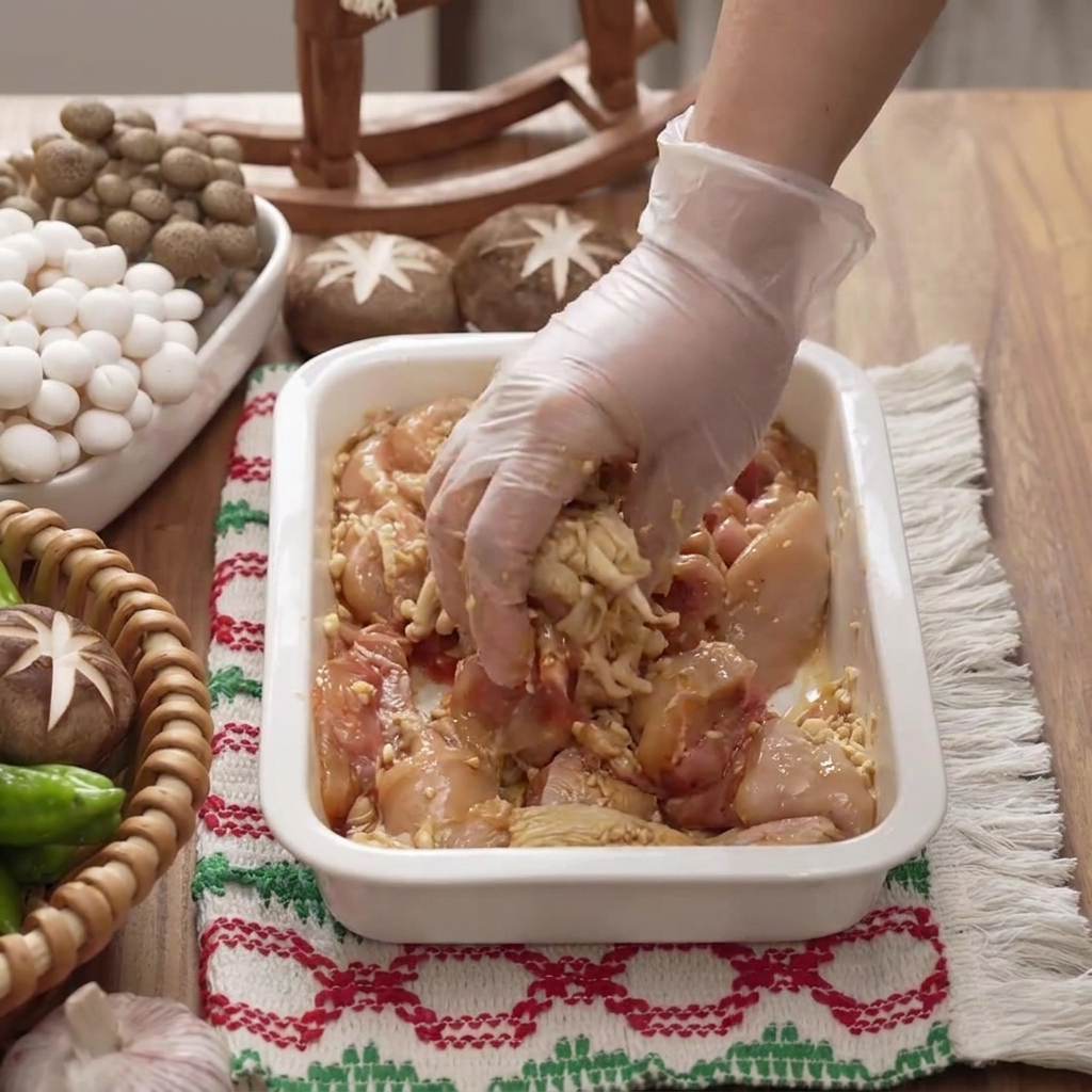 Une main gantée mélangeant et massant la marinade dans les morceaux de poulet à lintérieur dun plat blanc.