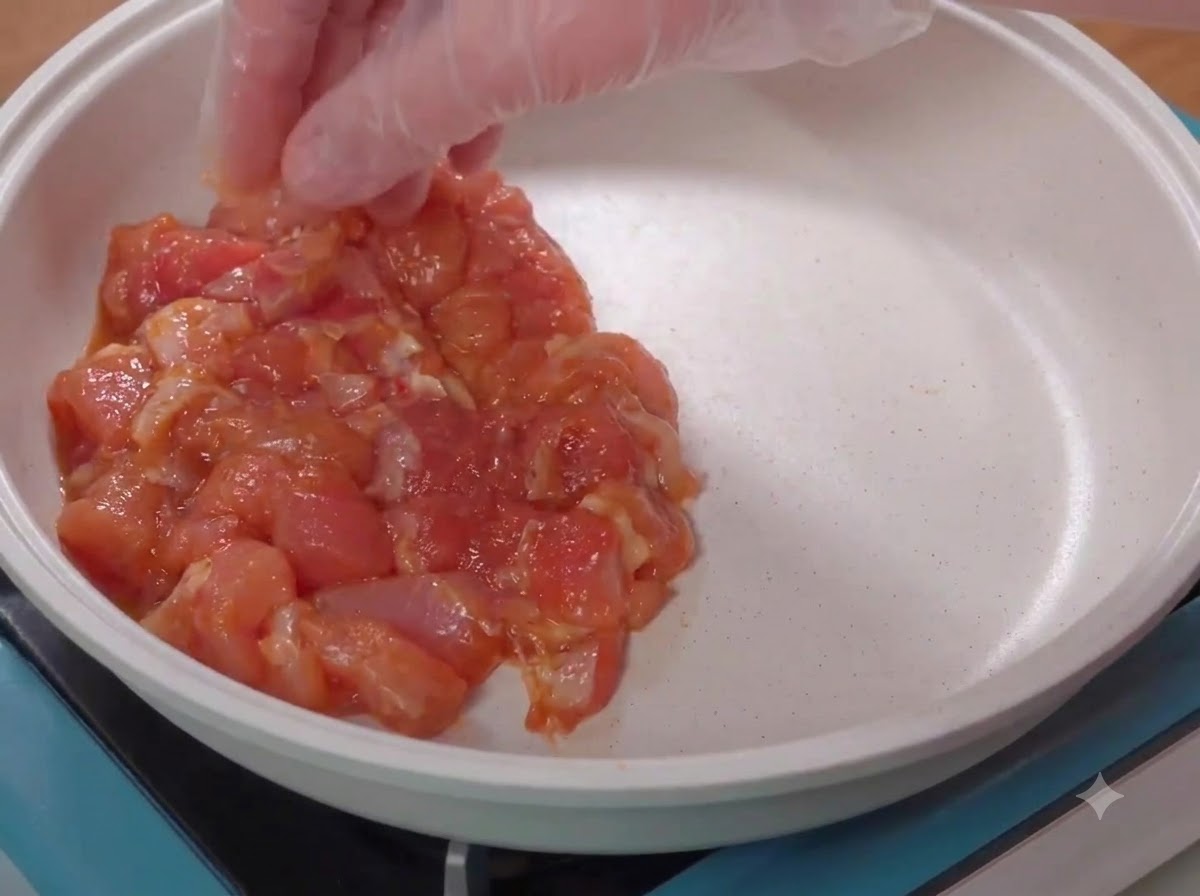 Une main gantée plaçant des morceaux de poulet cru marinés dans une poêle blanche.