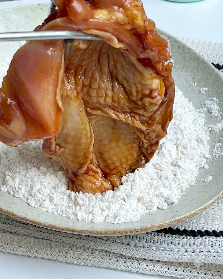 Des pinces tenant un morceau de poulet mariné au-dessus dune assiette blanche remplie de poudre de fécule de maïs blanche.