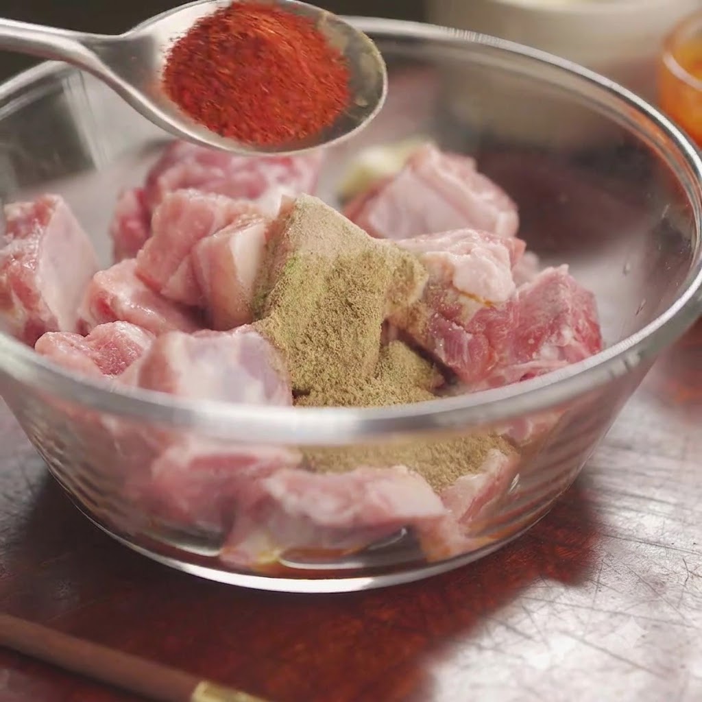 Poudre de piment rouge sur une cuillère tenue au-dessus dun bol de travers de porc crus recouverts dépices et dherbes mélangées.