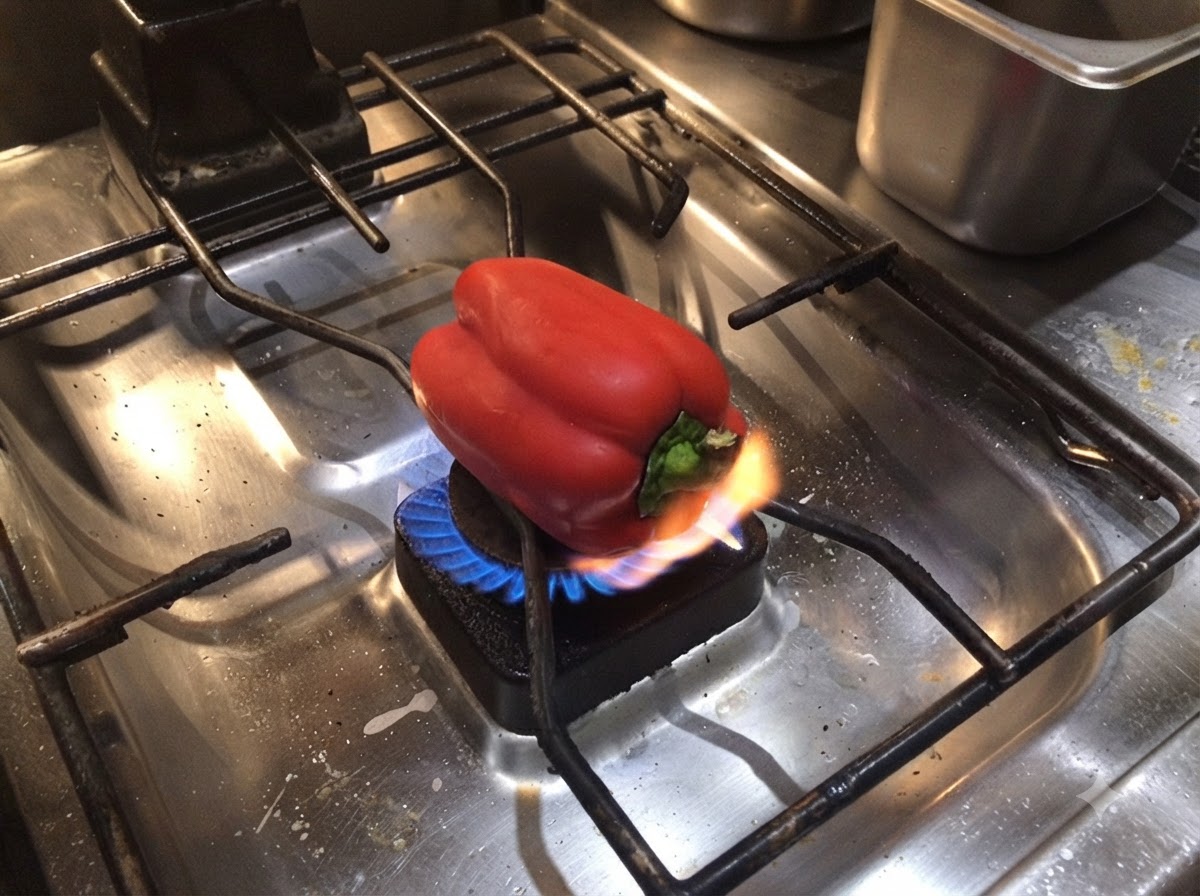 Un poivron rouge posé sur un brûleur de cuisinière à gaz avec une flamme nue noircissant sa base.