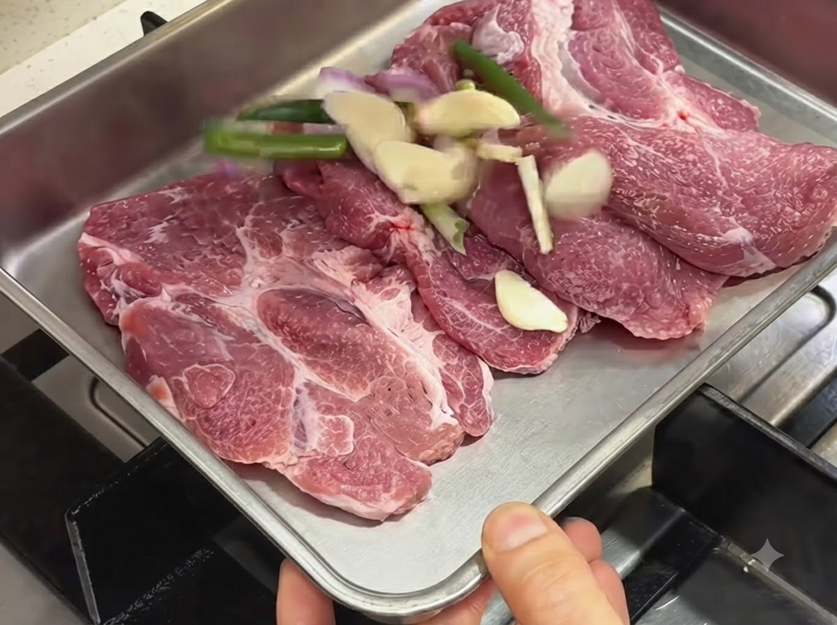 Côtes de porc crues disposées dans un plat en métal aux côtés dail, doignons verts et déchalotes tranchés.