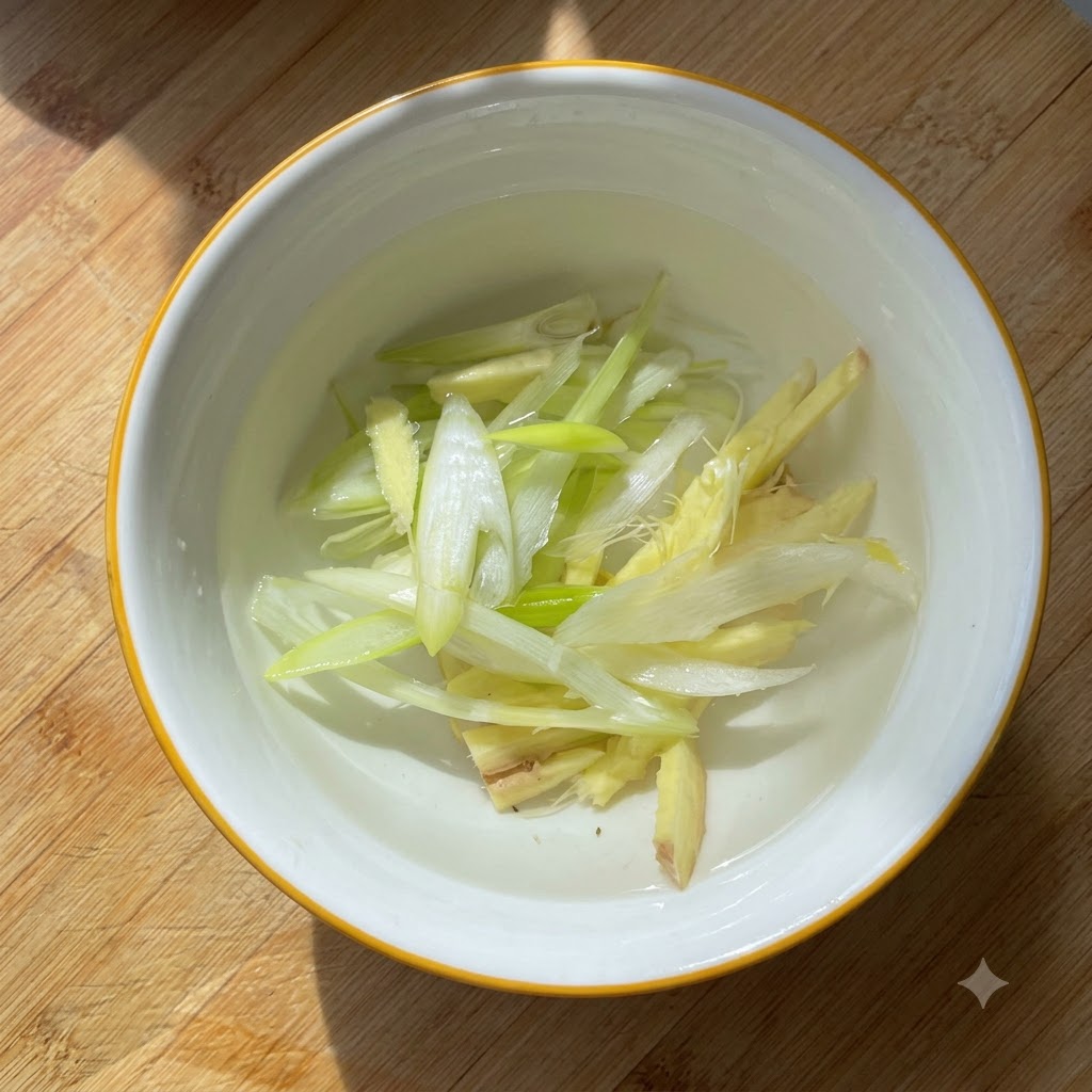 Tranches doignons verts et morceaux de gingembre infusant dans de leau à lintérieur dun petit bol blanc.