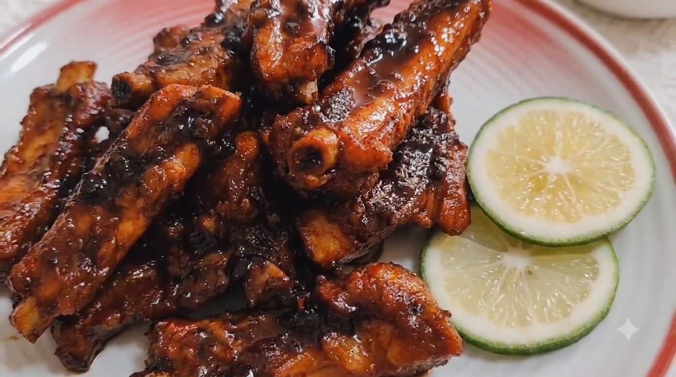 Travers de porc brillants braisés au café servis sur un plat en céramique à bordure rouge et garnis de tranches de citron vert frais.