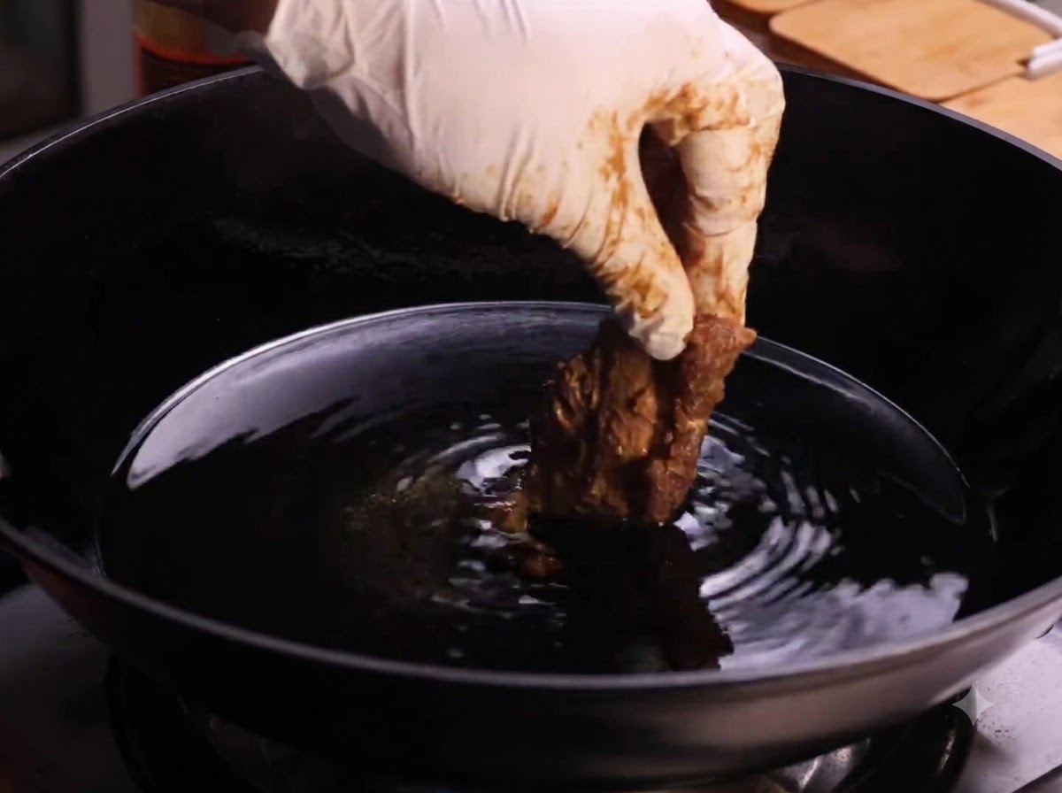Une main gantée abaissant un morceau de travers de porc enrobé dans un wok rempli dhuile de cuisson chaude.