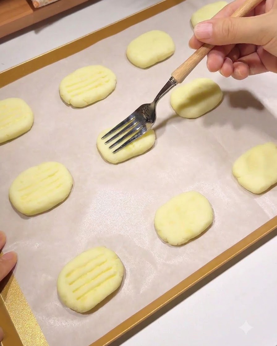 Une fourchette à manche en bois pressant un motif de lignes parallèles sur une galette de pommes de terre façonnée sur une plaque de cuisson recouverte de papier sulfurisé.