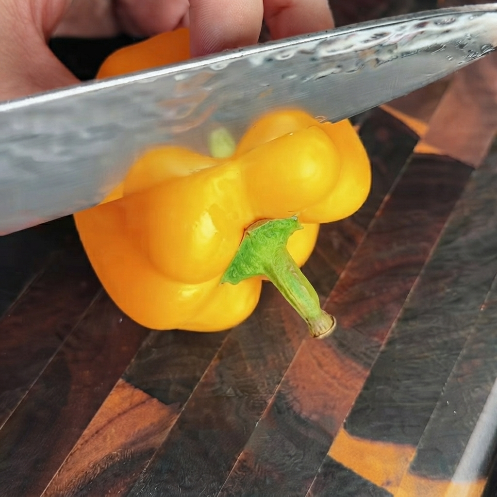 Un couteau bien aiguisé coupant la section supérieure dun poivron jaune vif sur une planche à découper en bois.
