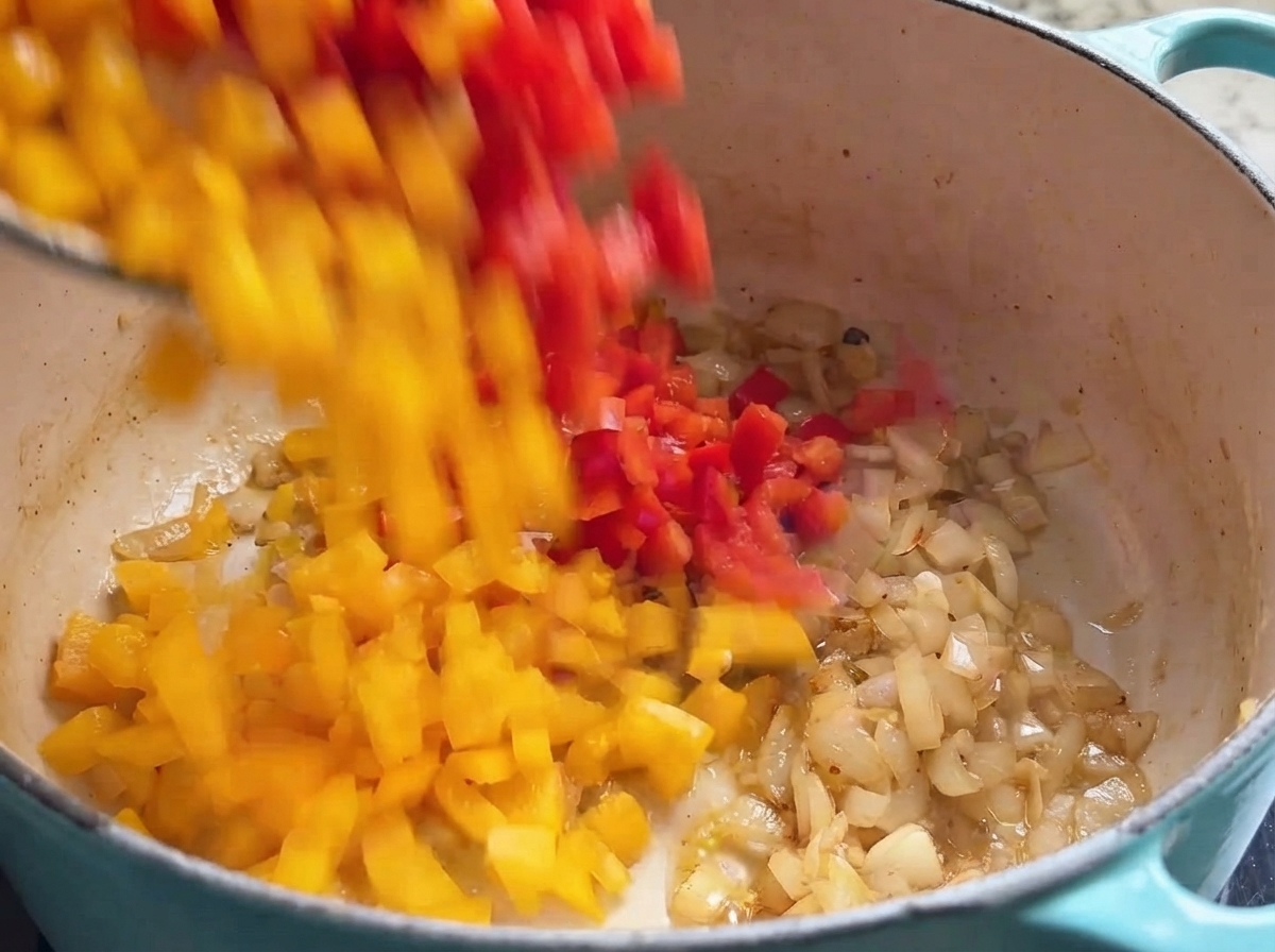 Des dés de poivrons rouges et jaunes tombant dans une casserole remplie doignons revenus.