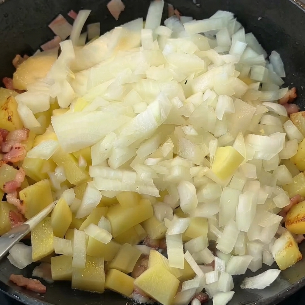 Des oignons en dés versés sur des pommes de terre et du bacon cuits dans une poêle.