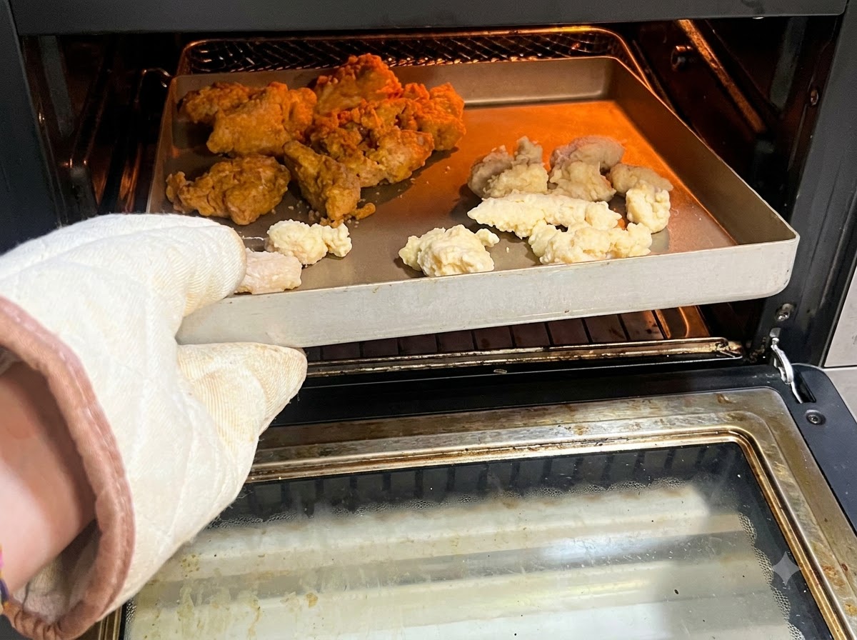 Un gant de cuisine tenant un plateau rempli de popcorn de poulet et dailes épicées crus ou surgelés à lintérieur dune friteuse sans huile.