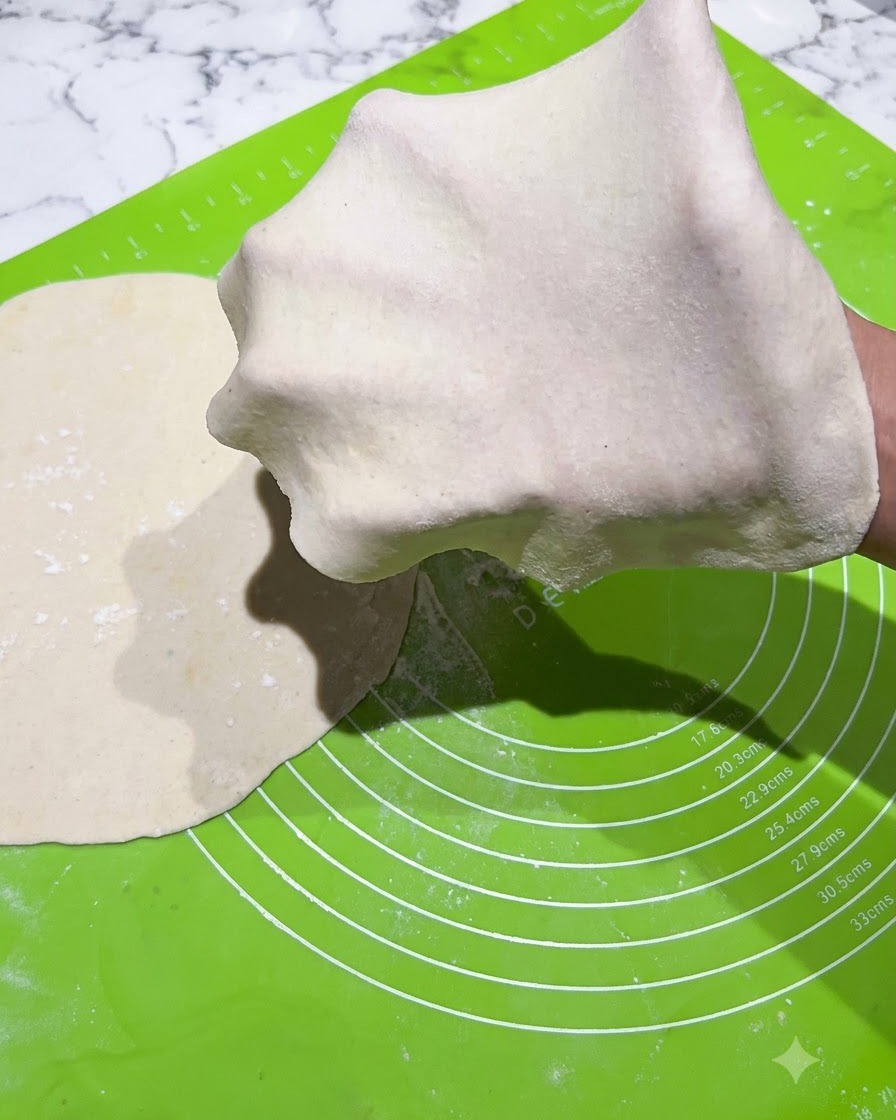 Une fine feuille de pâte étalée et étirée sur une main gantée sur un tapis vert.
