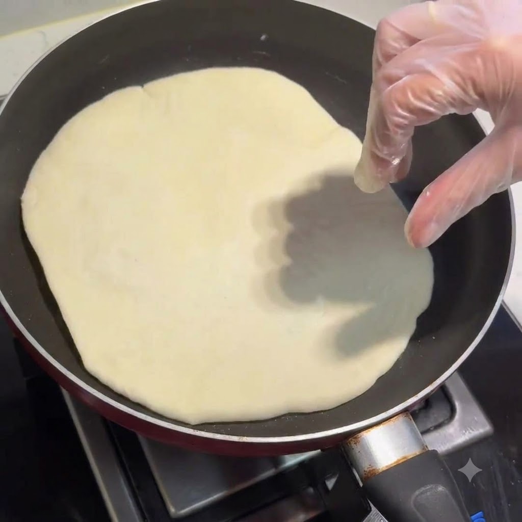 Une galette cuisant dans une poêle à frire avec une main gantée qui lajuste.