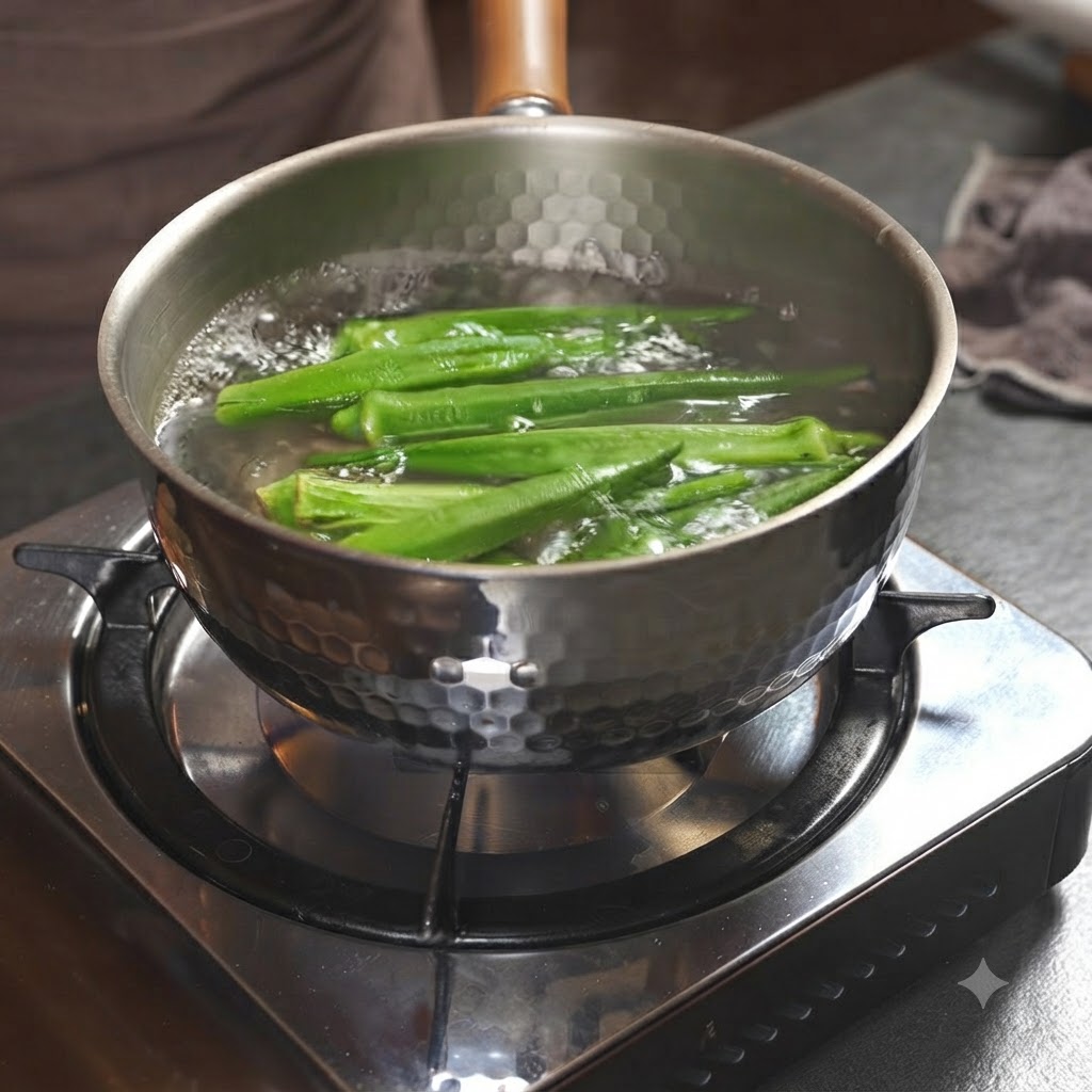 Gousses de gombos verts bouillant à lintérieur dune casserole métallique remplie deau sur un feu de cuisinière.