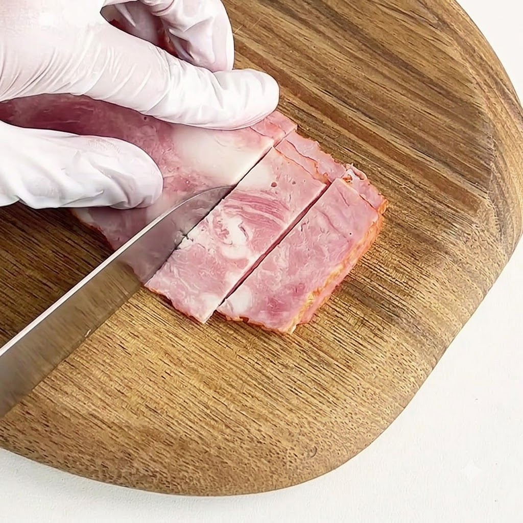 Une main gantée coupant du bacon en petits morceaux sur une planche à découper ronde en bois.