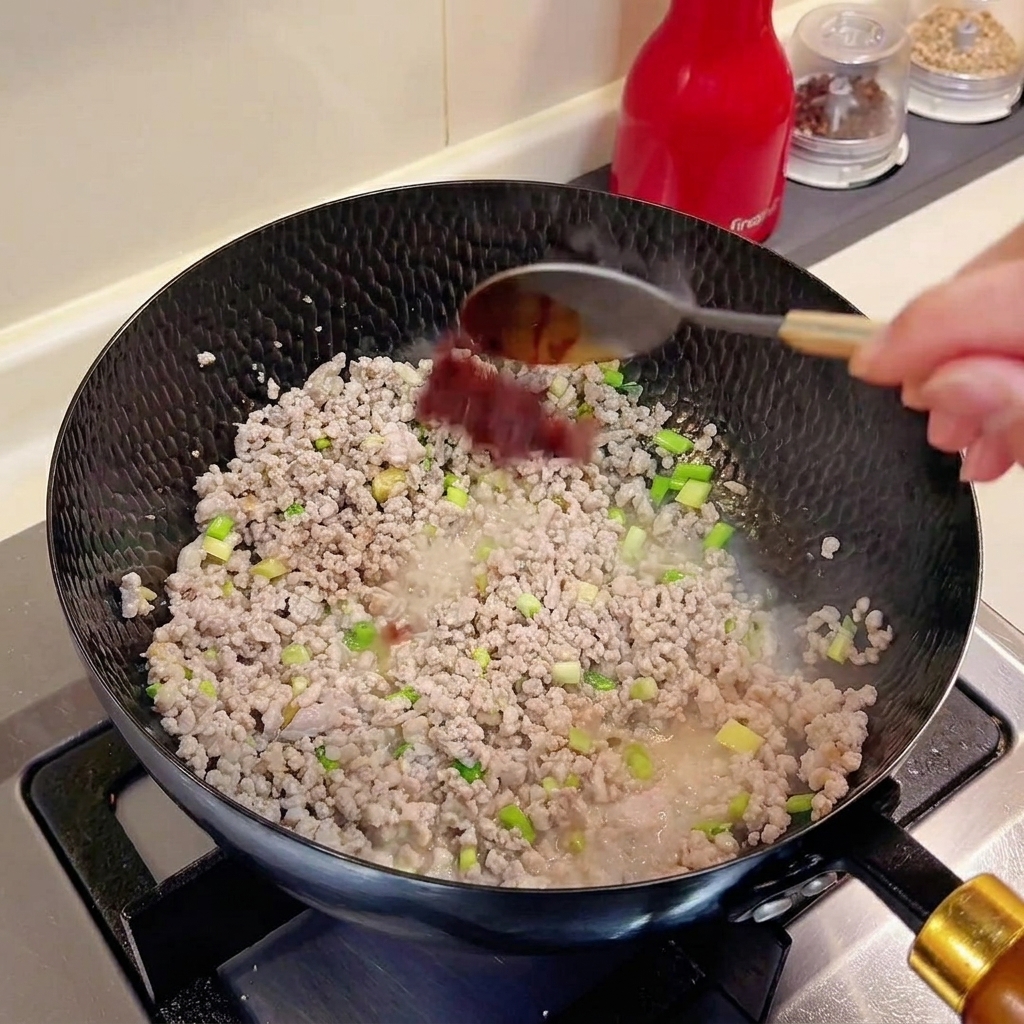Une cuillère de doubanjiang ajoutée dans un wok contenant la viande hachée et les aromates.