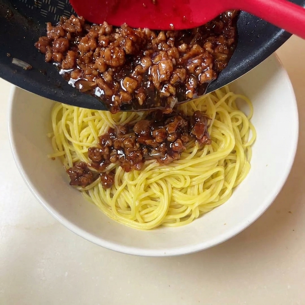 Une spatule rouge versant une sauce de viande hachée riche et foncée sur un bol de nouilles jaunes.