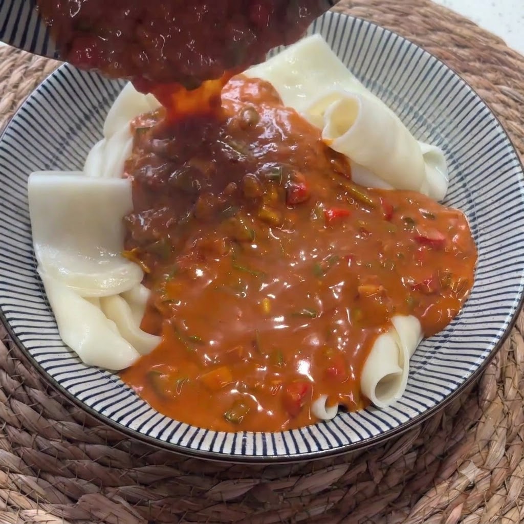 Sauce rouge riche versée dune louche sur un bol rempli de nouilles larges repliées.