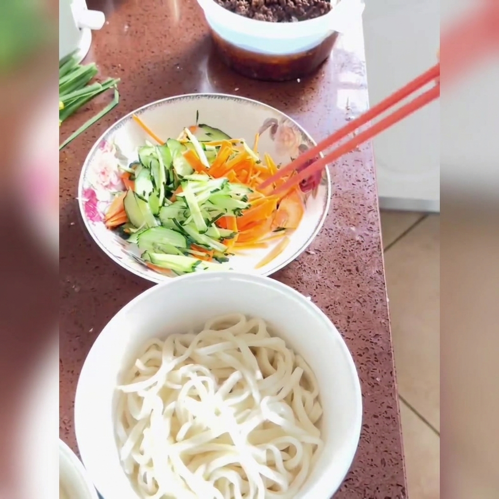 Sauce frite versée sur des nouilles fraîchement cuites garnies de concombre et de carottes râpés dans un bol blanc.
