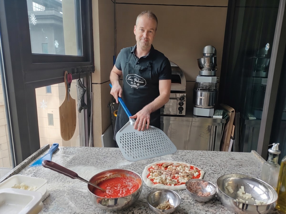 Un pizzaïolo tenant une grande pelle à pizza en métal perforé positionnée juste à côté dune pizza crue entièrement assemblée.