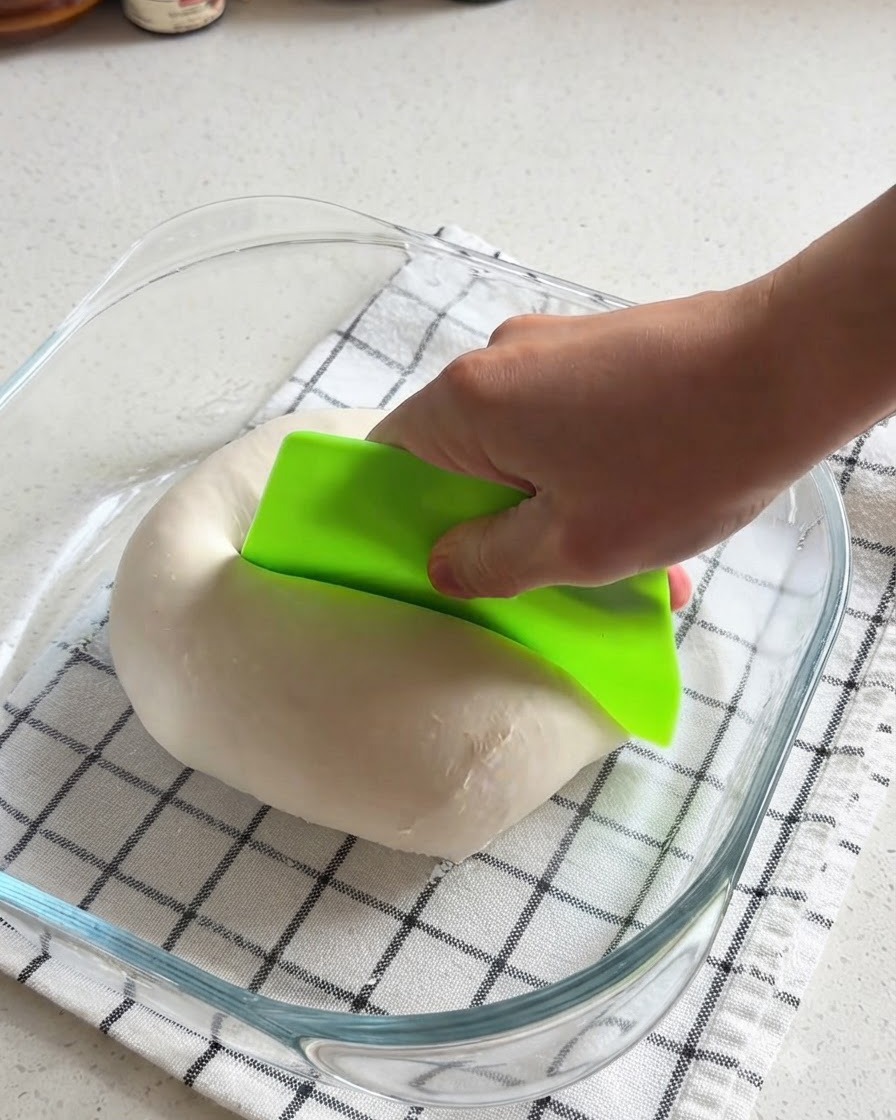 Une main utilisant un racloir de boulanger vert vif pour couper une boule de pâte à pizza lisse en deux dans un plat en verre.