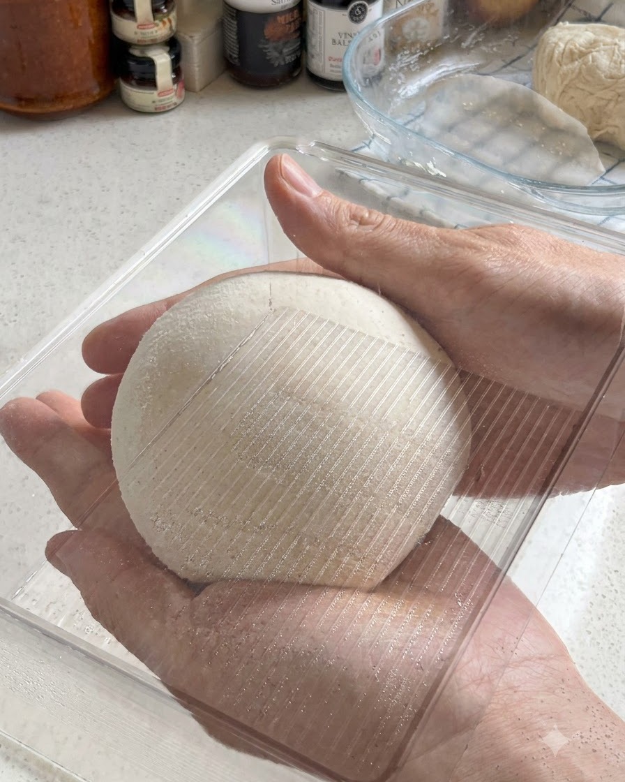 Deux mains tenant une boule de pâte à pizza parfaitement arrondie alors quelle est placée dans un récipient de fermentation en plastique transparent.