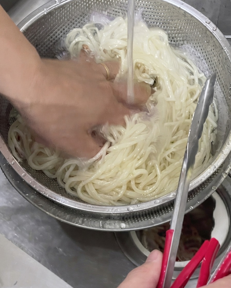 Une main remuant des nouilles udon cuites dans une passoire en métal sous leau courante froide.