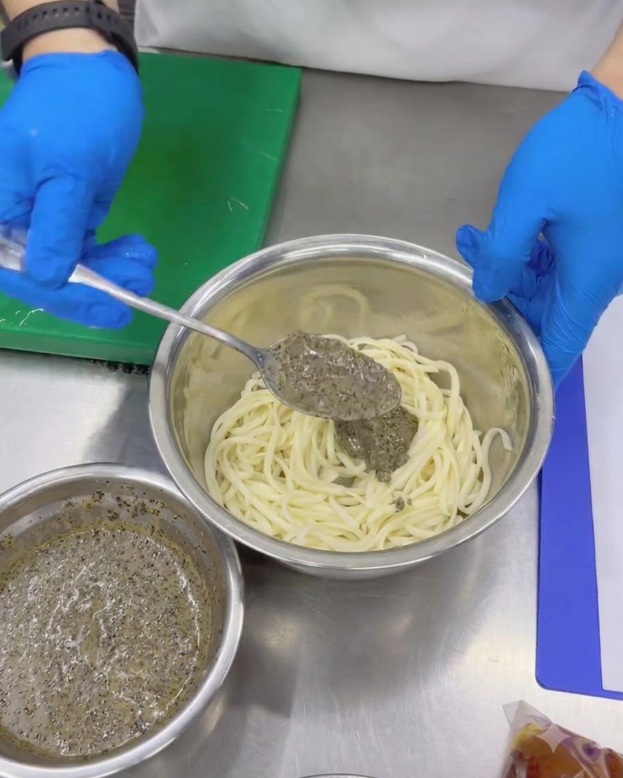Mains gantées utilisant une cuillère pour ajouter une épaisse sauce à la truffe noire aux nouilles udon froides dans un bol à mélanger en métal.