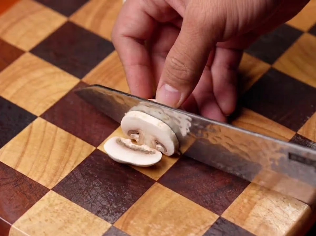 Champignons de Paris tranchés sur une planche à découper en bois à motifs vichy, puis versés dans une poêle noire.