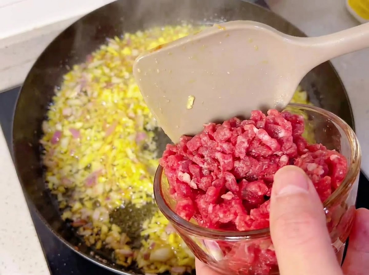 Un petit bol en verre versant des cubes de bœuf dans une poêle chaude remplie doignons et dail sautés.