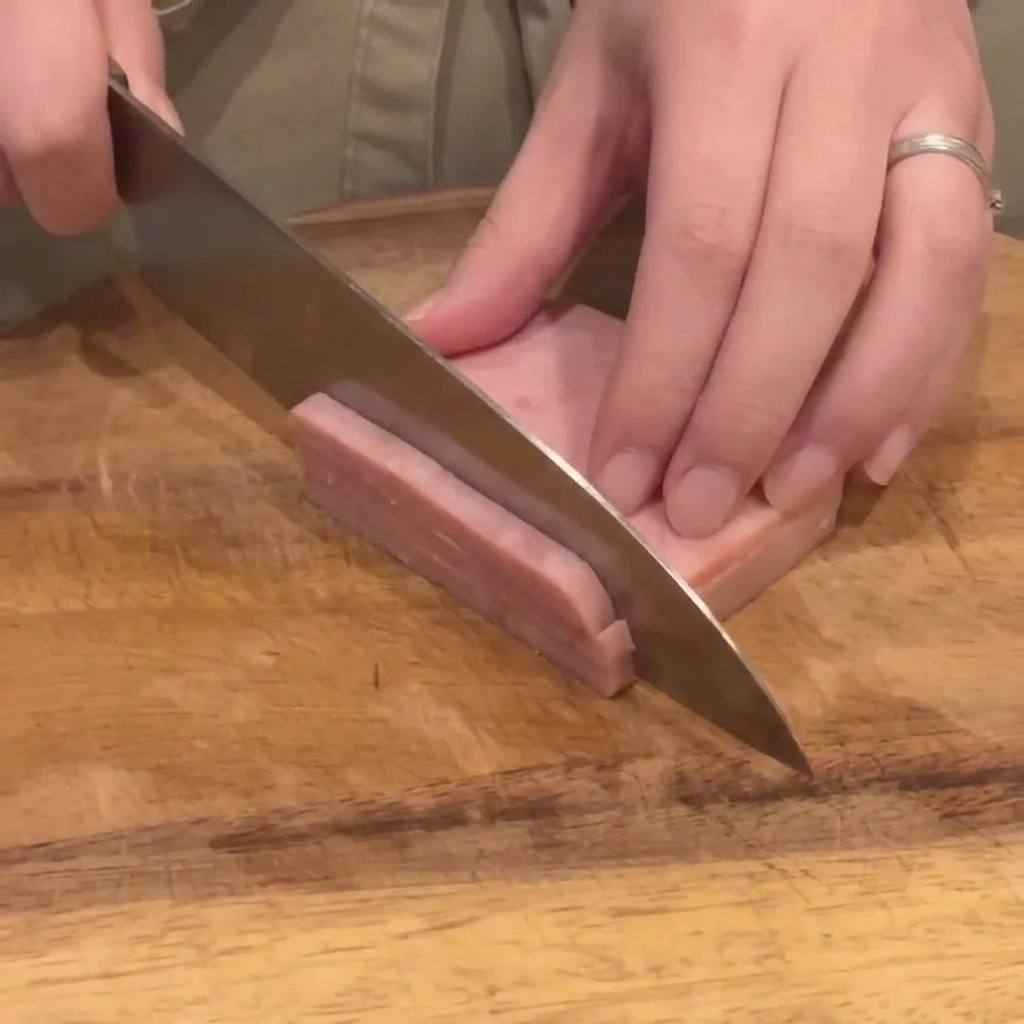 Un couteau de cuisine tranchant un bloc de jambon rose en lanieres fines et regulieres sur une planche en bois.