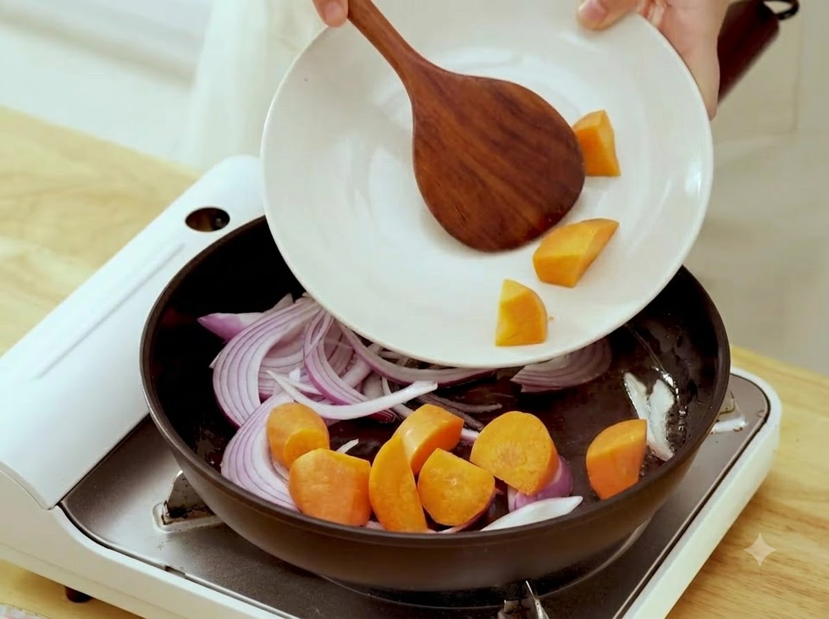 Des morceaux de carottes crues ajoutés depuis une assiette dans une poêle contenant des oignons sautés.