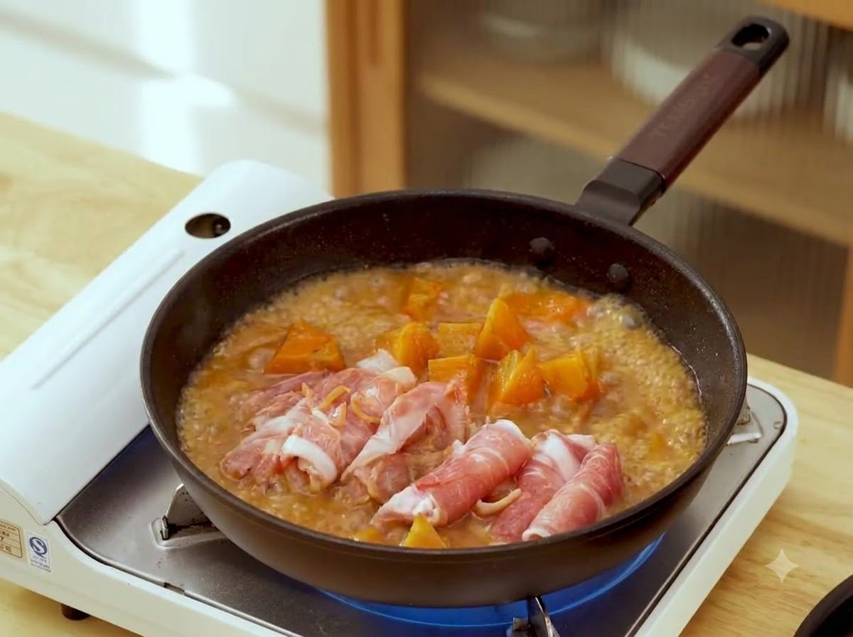 Des tranches de bœuf crues enroulées disposées à lintérieur dune poêle avec un bouillon de miso brun frémissant et des morceaux de carotte.