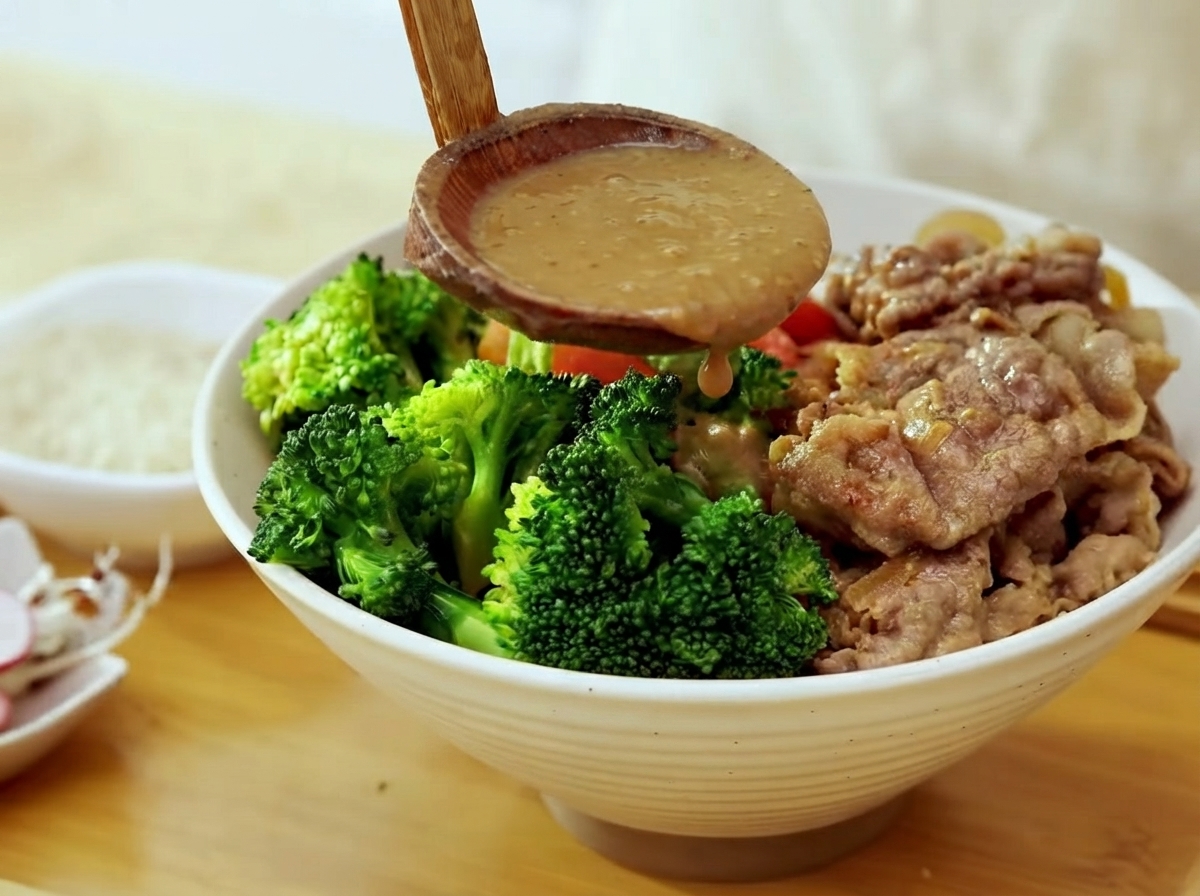 Une louche en bois versant une sauce miso brune épaisse sur un bol rempli de brocoli, de bœuf cuit et de riz blanc.