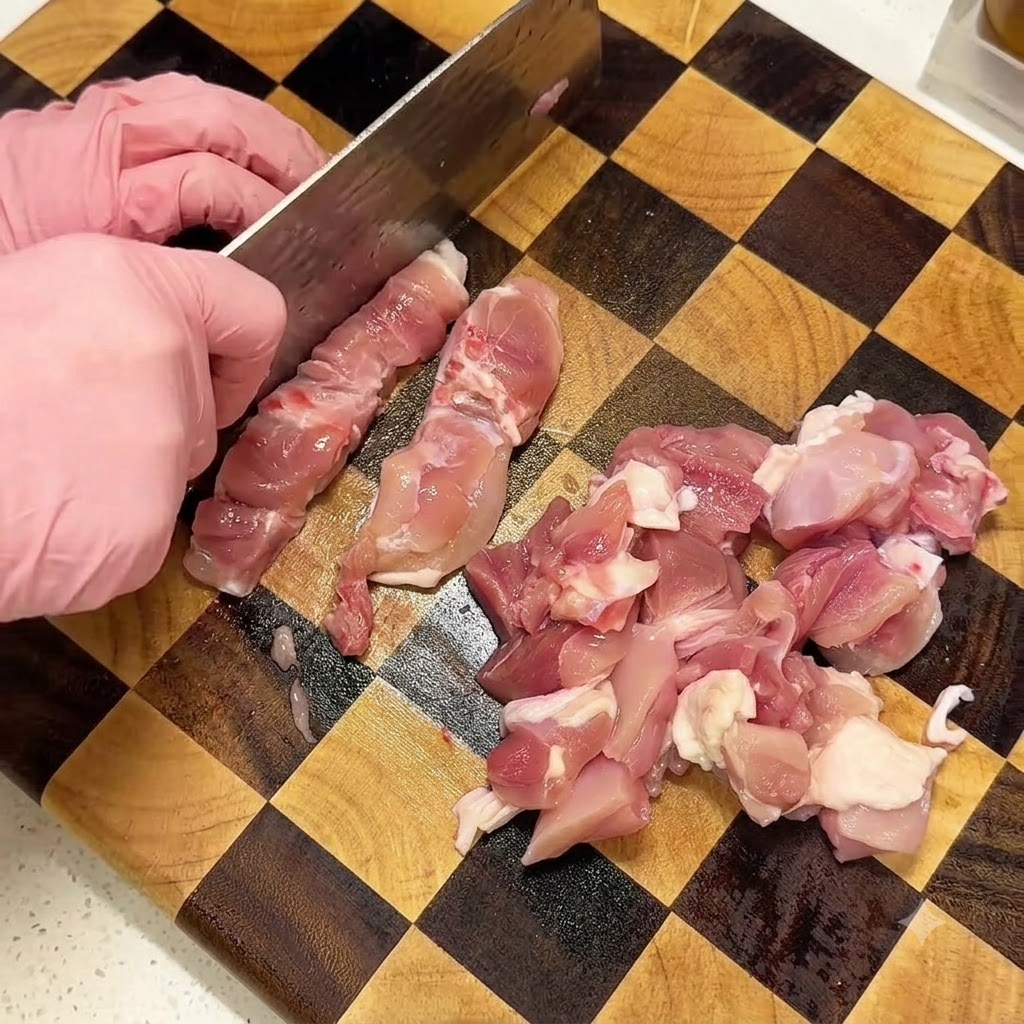 Mains portant des gants roses utilisant un couperet pour découper des cuisses de poulet crues sur une planche à découper en bois à damier.