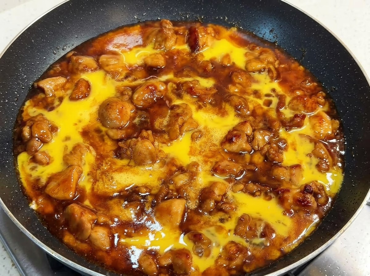 Une poêle remplie de morceaux de poulet et dune sauce riche surmontée de liquide dœuf battu cru, couverte dun couvercle.