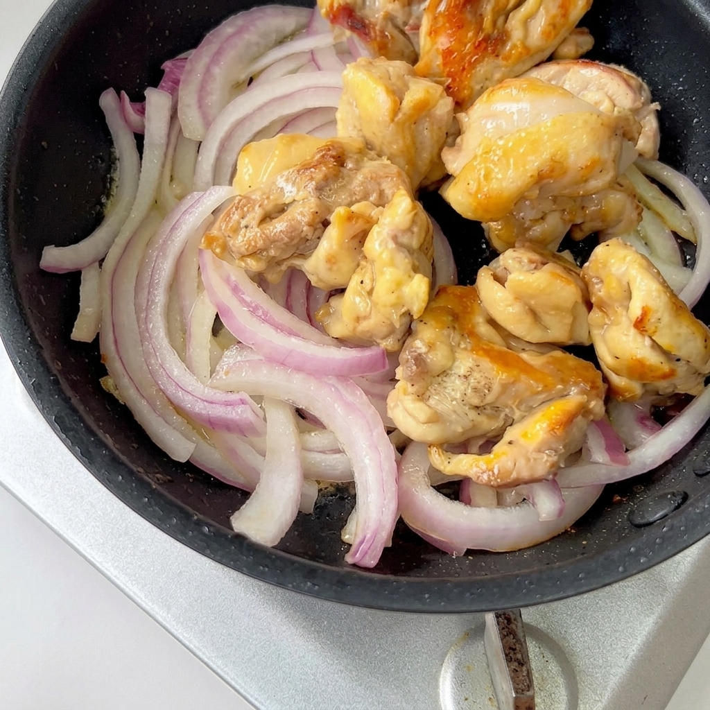 Morceaux de poulet cuits et oignons rouges émincés mijotant dans une poêle avec de la sauce.