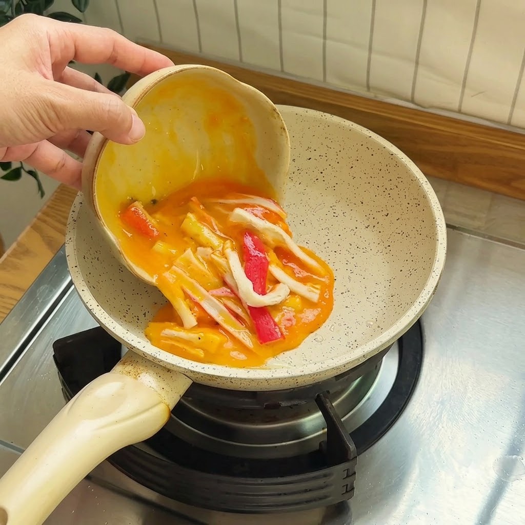 Des œufs battus mélangés à des morceaux de surimi rouges et blancs versés dans une petite poêle antiadhésive.