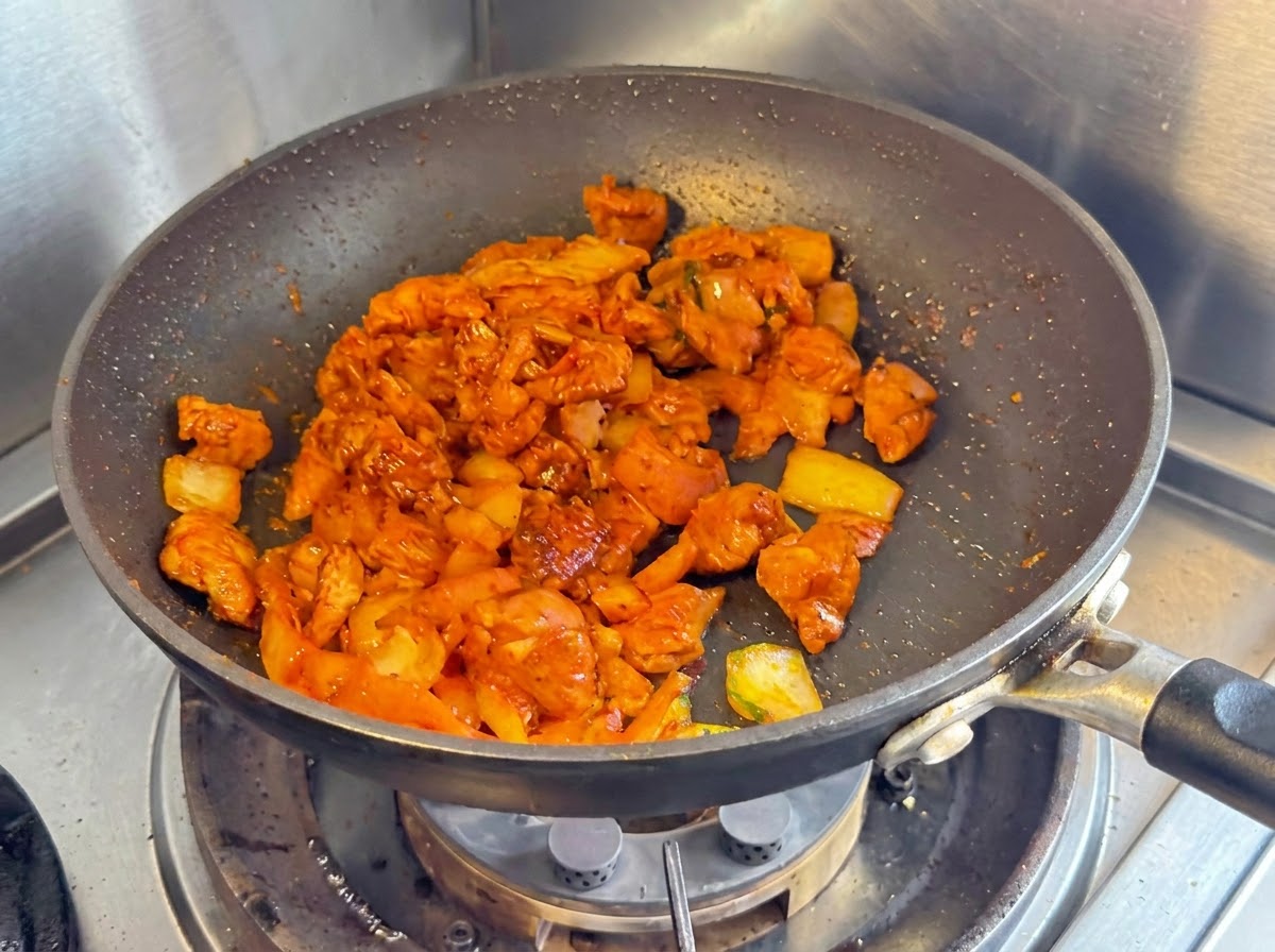Dés de blanc de poulet, oignons et kimchi haché sautés ensemble dans une poêle antiadhésive sombre.