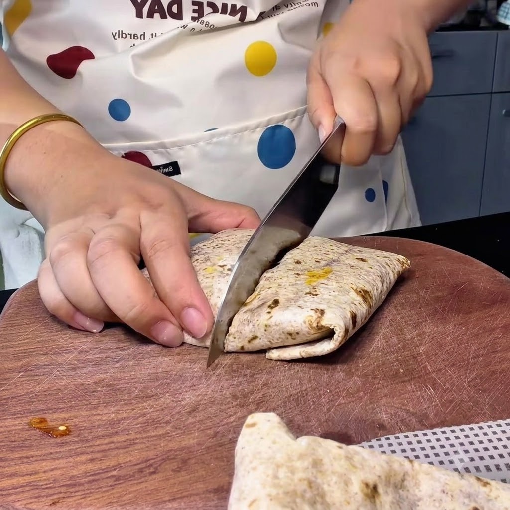Une personne utilisant un grand couteau de chef pour couper en diagonale un wrap au blé complet sur une planche en bois.