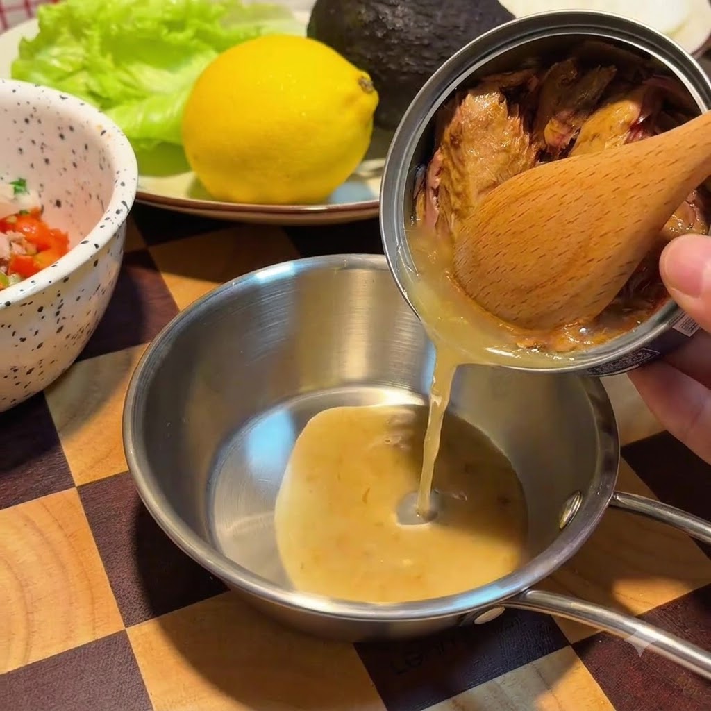 Une cuillère en bois retient des morceaux de thon à lintérieur dune boîte en métal tandis quune huile dorée sécoule dans un petit bol en acier inoxydable.