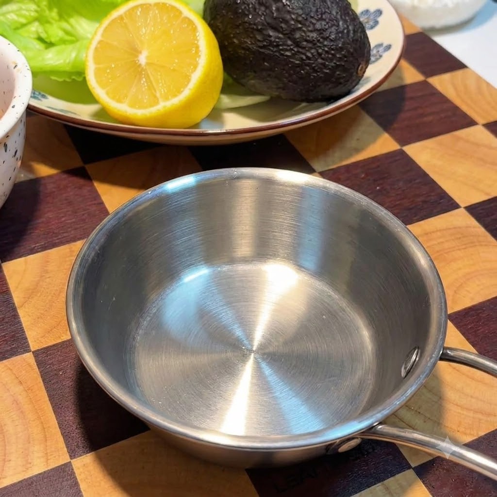 Un petit bol vide en acier inoxydable posé sur une planche en bois à carreaux à côté dune assiette avec un citron et un avocat.